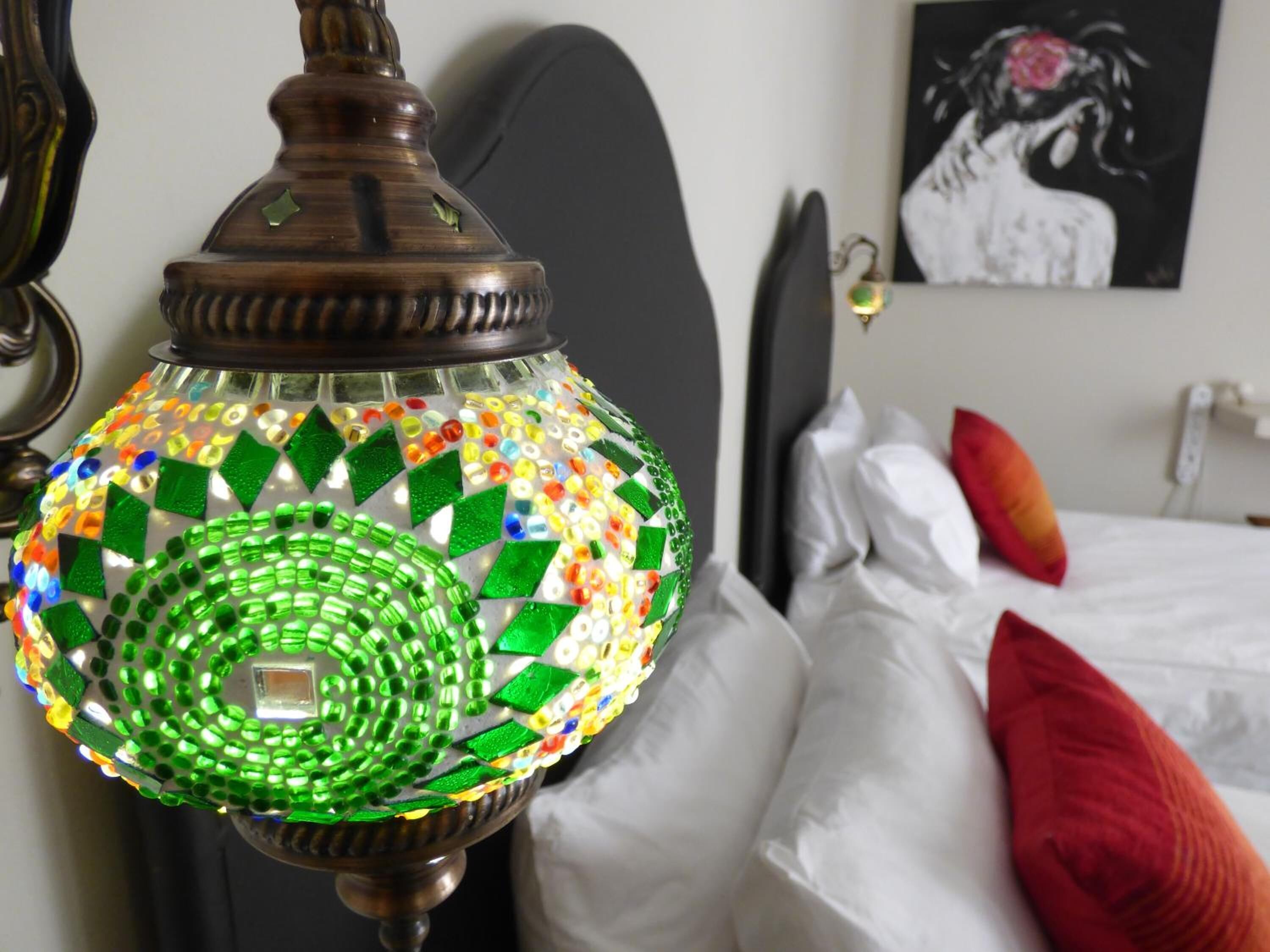 Una habitación con cama, almohadas, un cuadro en la pared y una lámpara decorativa con diseño de mosaico.