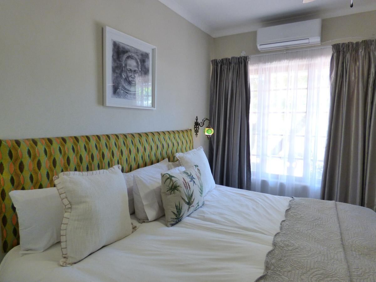 Un lit bien fait, avec du linge de lit blanc et des coussins décoratifs, une tête de lit à motifs, une œuvre d’art encadrée au mur et une fenêtre avec des rideaux.