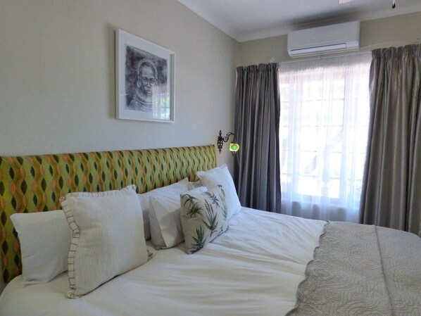 A neatly made bed with white linens and decorative pillows, a patterned headboard, a framed artwork on the wall, and a window with curtains. - Villa Africa Guesthouse (Tsumeb)