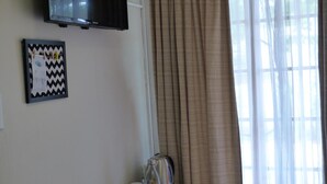 Una habitación con una televisión de pantalla plana montada en la pared, un cuadro con un diseño en zigzag, una mesita con una tetera, tazas y una botella de vidrio, y una ventana con cortinas translúcidas.