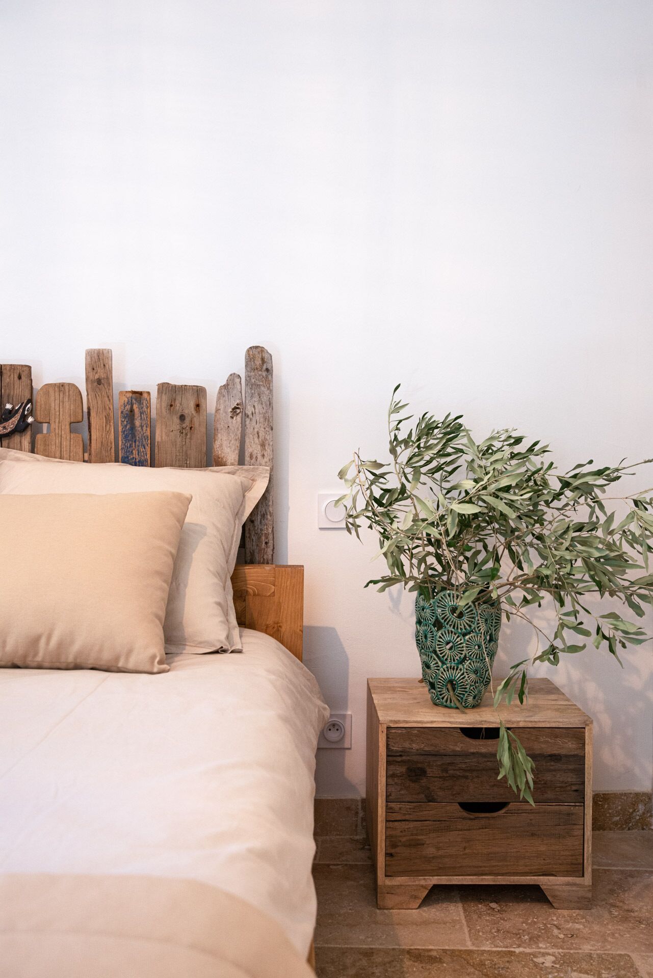 Ein Schlafzimmer mit einem hölzernen Kopfteil, einem Bett mit beigefarbener Bettwäsche und einem Nachttisch mit einer vase mit grünem Muster und einer Pflanze.