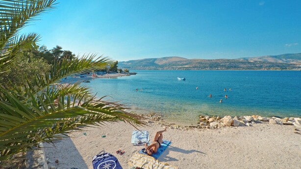 A beach with clear blue water, a sandy shore, and a person lying on a towel.