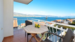 A balcony with a wooden table and wicker chairs, overlooking a coastal view with buildings and mountains in the distance.