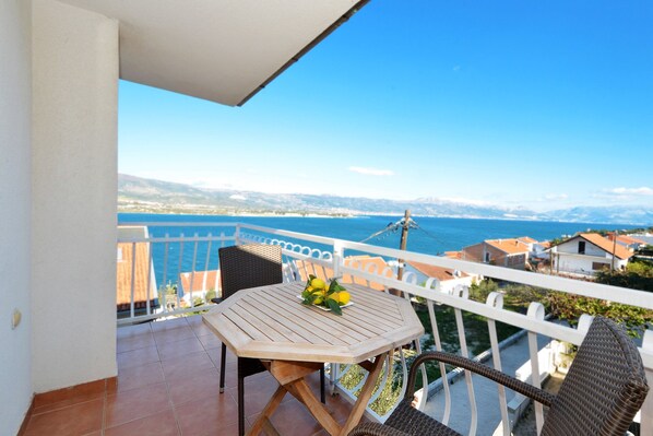 A balcony with a wooden table and wicker chairs, overlooking a coastal view with buildings and mountains in the distance.