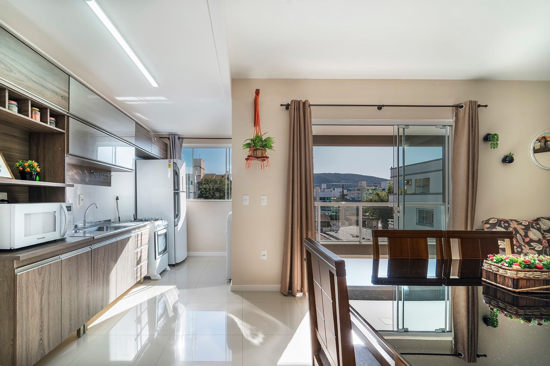 Een moderne keuken met een koelkast, fornuis en magnetron. Er is een eethoek met een tafel en stoelen. De kamer heeft grote ramen met gordijnen en uitzicht op gebouwen.