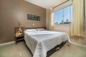 A neatly made bed in a room with a large window, a painting on the wall, and a nightstand with a lamp. - Aluguel Apartamento 2 quartos sendo 1 suíte - 522 (Bombinhas)