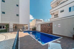 A swimming pool with a wooden deck, a brick wall, and a white building in the background. - Aluguel Apartamento 2 quartos sendo 1 suíte - 522 (Bombinhas)