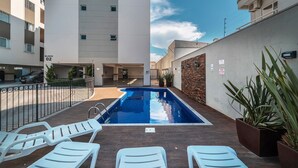 A swimming pool with lounge chairs, a brick wall, and a building in the background. - Aluguel Apartamento 2 quartos sendo 1 suíte - 522 (Bombinhas)