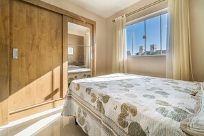 A bedroom with a bed, a wardrobe, a window with curtains, and a view of buildings. - Aluguel Apartamento 2 quartos sendo 1 suíte - 522 (Bombinhas)