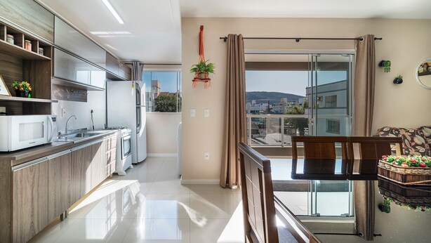A modern kitchen with a refrigerator, stove, and microwave. There is a dining area with a table and chairs. The room has large windows with curtains and a view of buildings outside. - Aluguel Apartamento 2 quartos sendo 1 suíte - 522 (Bombinhas)