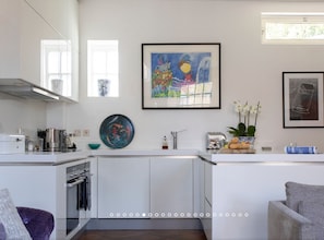 Une cuisine moderne avec des meubles blancs, une hotte de cuisinière en acier inoxydable, un grille-pain et une machine à café. Un tableau encadré est accroché au-dessus du comptoir, et une plante en pot est posée sur le comptoir.