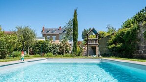 Una piscina amb un pont de fusta, envoltada de vegetació i una casa al fons.