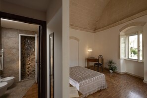 A bathroom with a shower and two sinks, a bedroom with a bed, a desk, a chair, and a window with a view. - Ai Terrazzini (Matera)