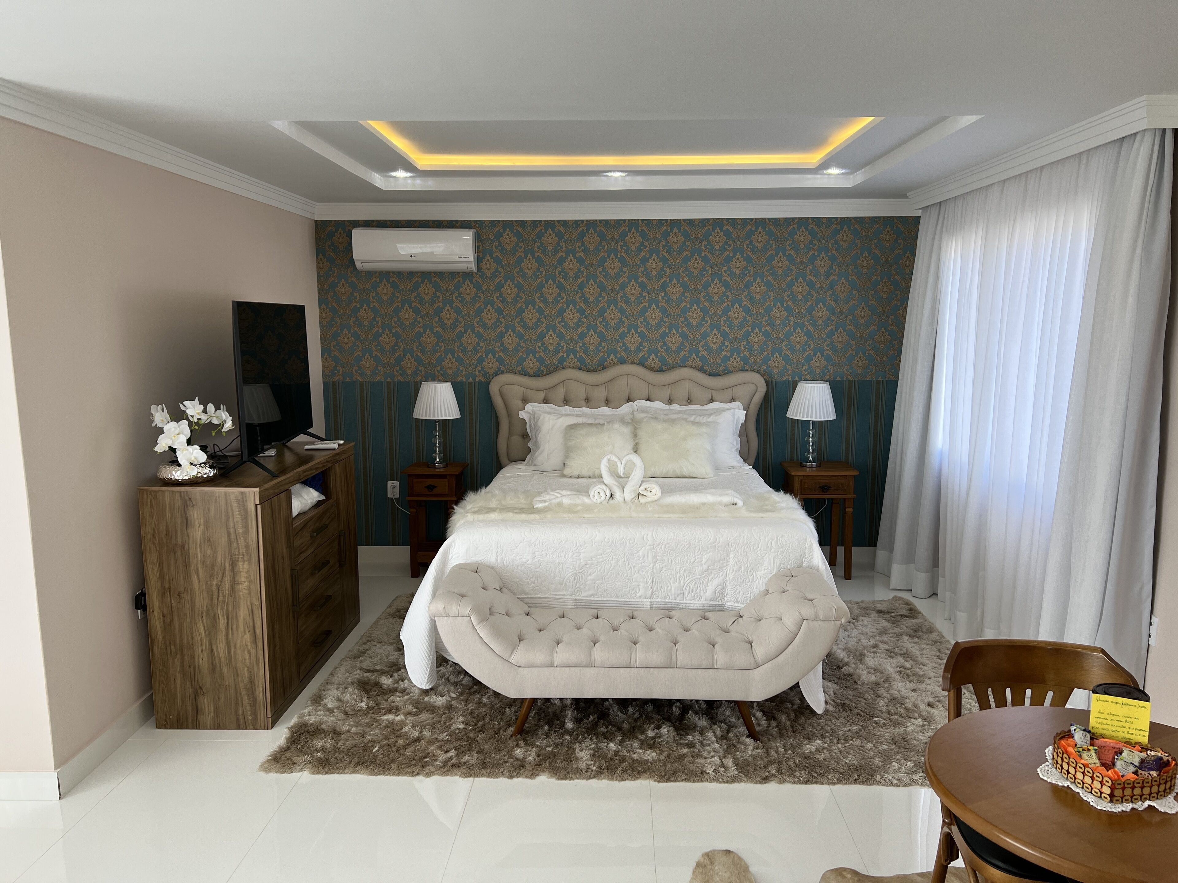 A bedroom with a tufted headboard bed, two bedside tables with lamps, a wooden dresser, a fluffy rug, and a dining table with a cake.