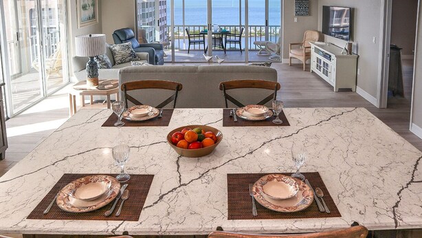 Un coin repas avec une table en marbre dressée pour quatre personnes, un saladier rempli de fruits et une vue sur l’océan grâce à de grandes fenêtres.