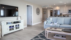 Un salon moderne doté d’un téléviseur à écran plat, d’un meuble de rangement blanc, d’un canapé bleu clair et d’un coin cuisine avec des appareils en acier inoxydable.