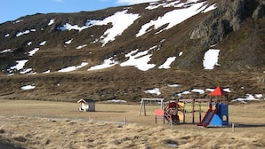 Une aire de jeux avec un toboggan et des structures d’escalade, un petit abri en bois et une zone délimitée par une clôture, recouverte d’herbe sèche.