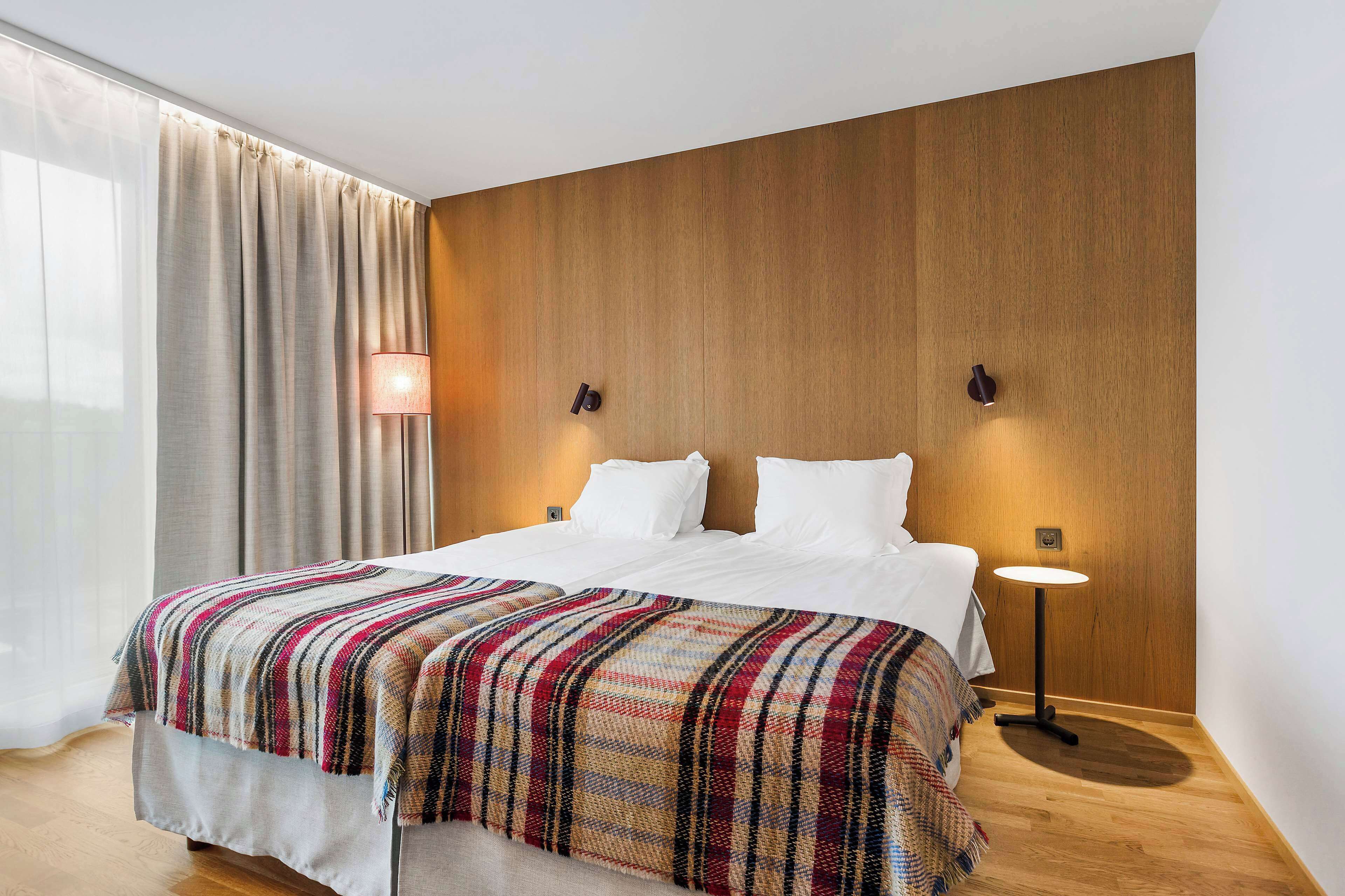 Ett hotellrum med en väggpanel i trä, en stor säng med ett rutigt överkast, ett sängbord med en lampa och utsikt över naturen genom tunna gardiner.