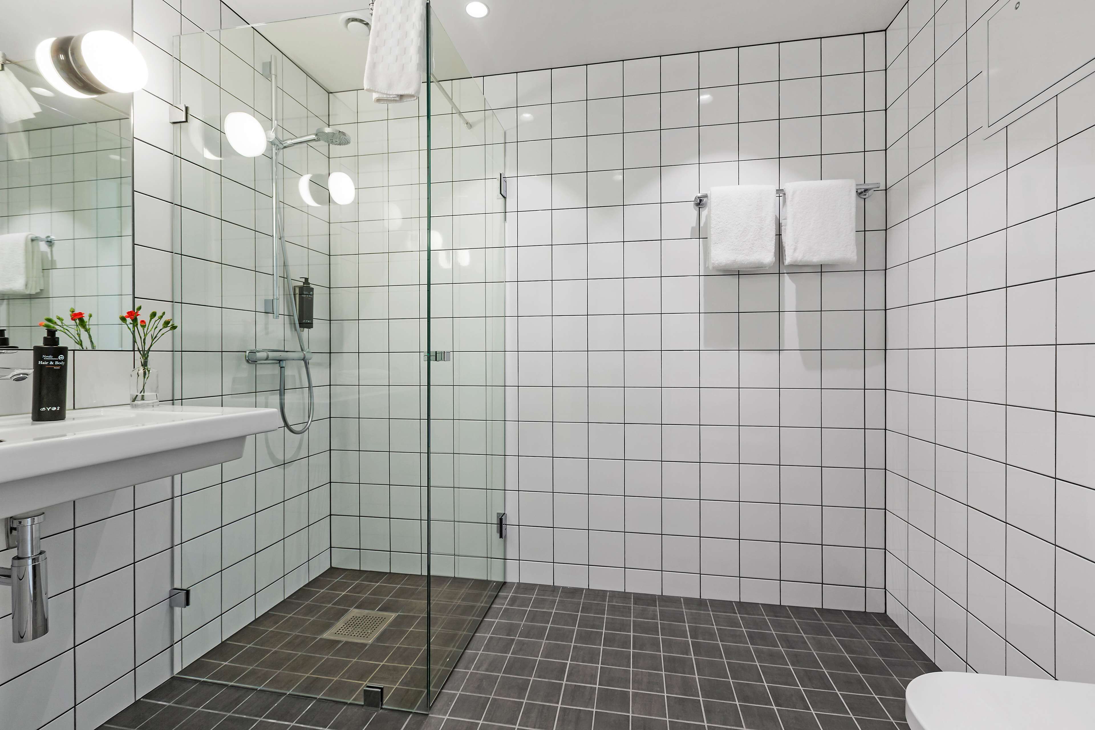 Ett modernt badrum med en duschavdelning med glasväggar, vita kakelväggar, en vit tvättställ med spegel och en toalett.