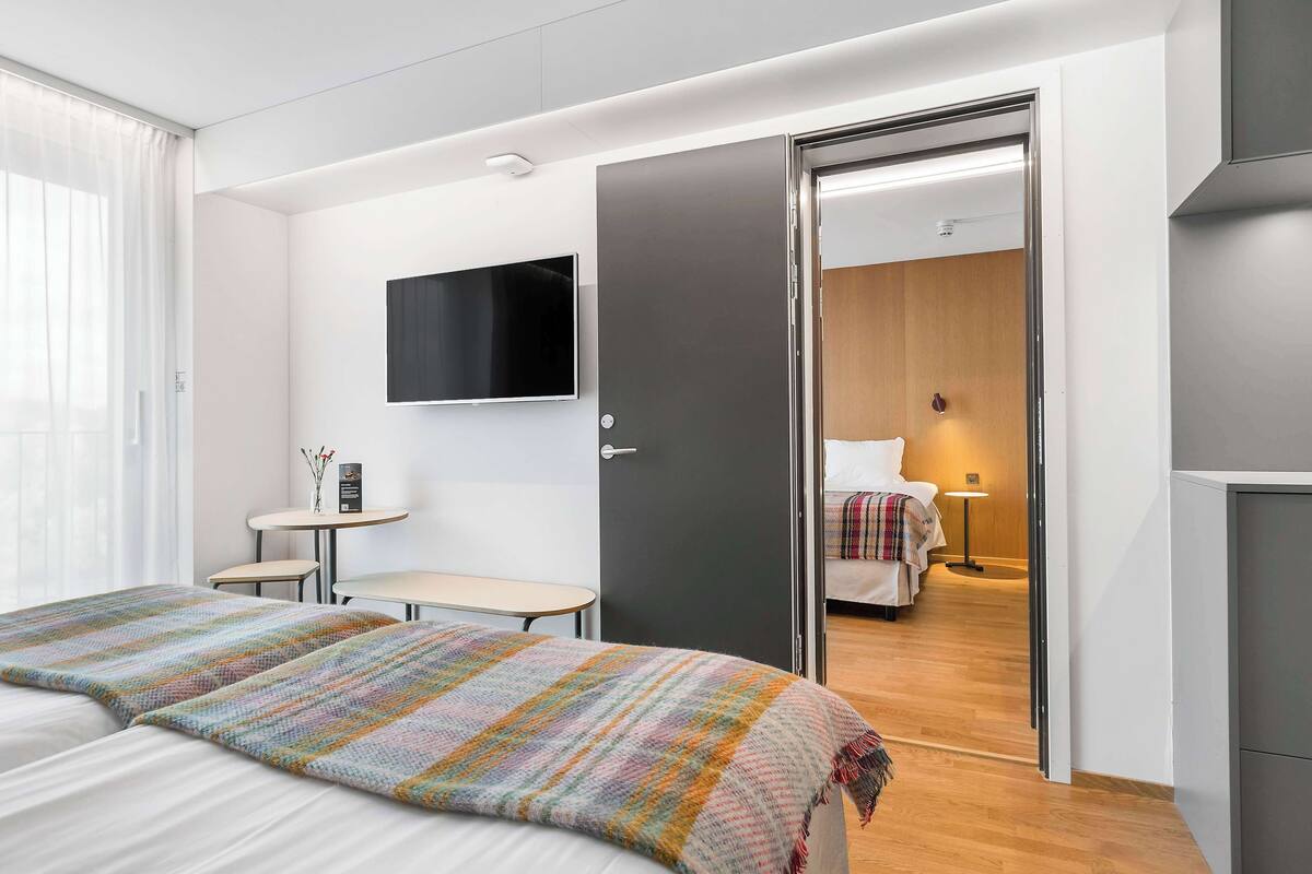 A modern hotel room with a large bed, a small table, a TV mounted on the wall, and a door leading to another room.