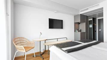 Una camera d'albergo moderna con un letto, un tavolino, una sedia in vimini, una TV a parete e un armadio.