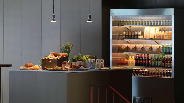 Una zona de bar con un refrigerador lleno de bebidas, una caja de madera con pasteles y luces colgantes.