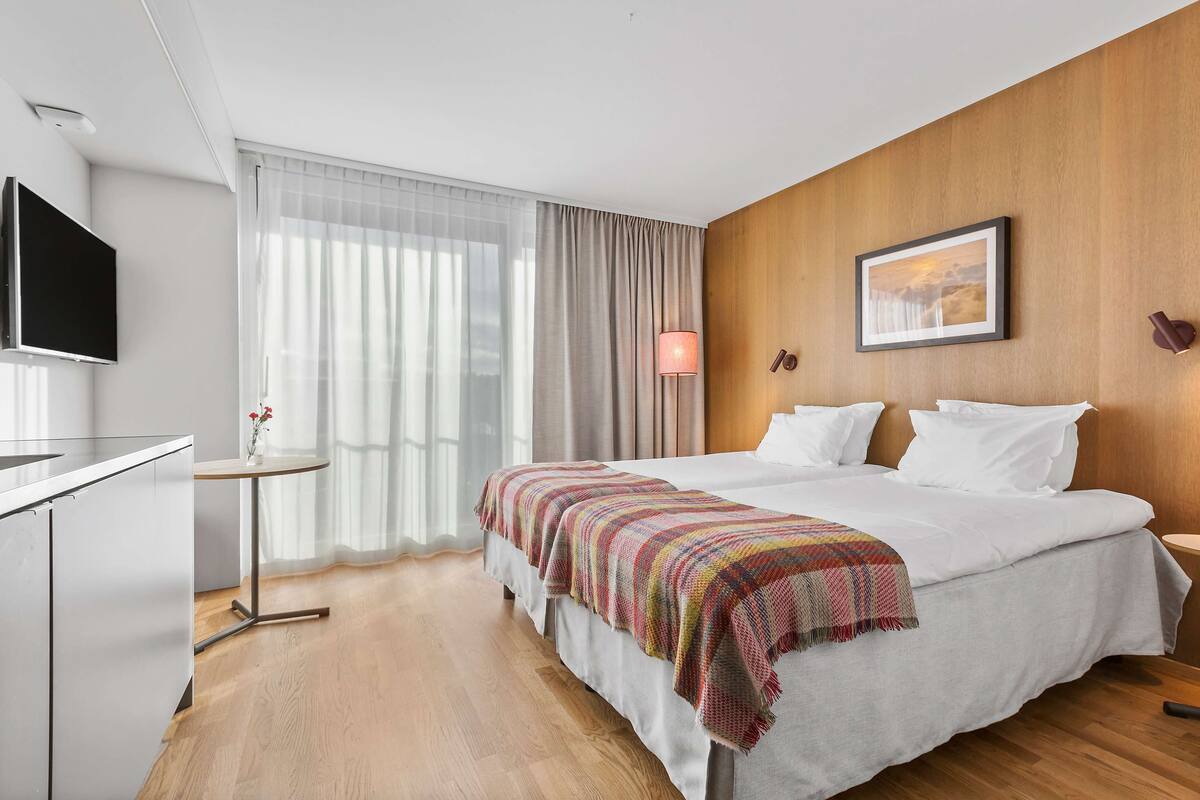A modern hotel room with a large bed, a TV, a small table, and a window with curtains.