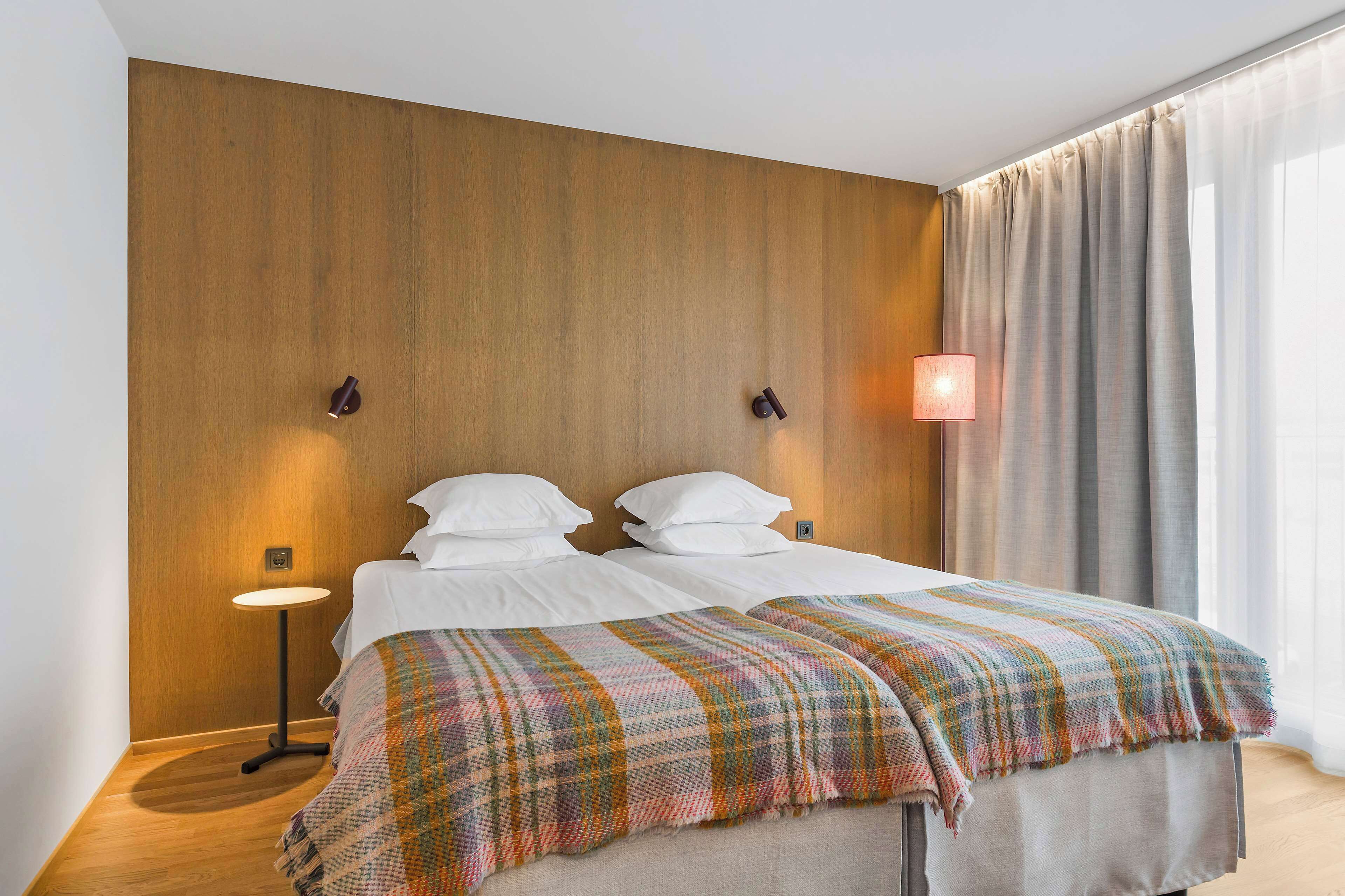 A hotel room with a wooden accent wall, two beds with white linens, a colorful plaid blanket, a small table lamp, and a large window with curtains.