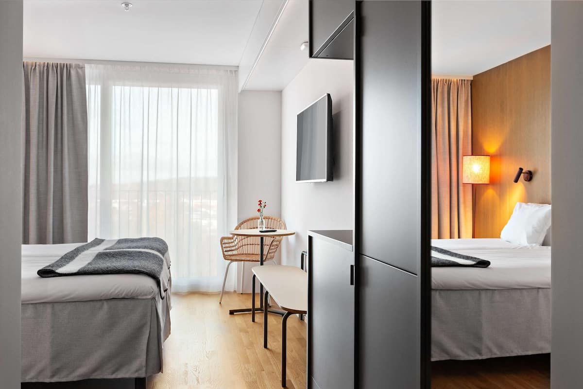A hotel room with two beds, a desk, a chair, a TV, and a lamp.