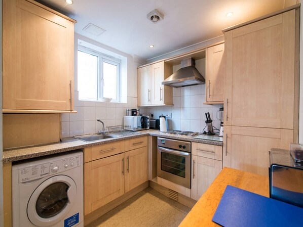 A compact kitchen with wooden cabinets, a built-in washing machine, a stove, and a window providing natural light.