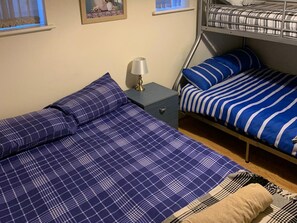 A bedroom with a bunk bed, a single bed with a plaid duvet, a bedside table with a lamp, and a framed picture on the wall.