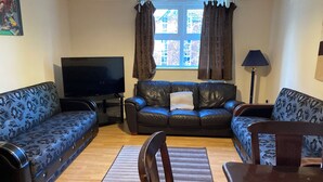 Ein Wohnzimmer mit einer Sofagarnitur, einem Fernseher, einer Lampe und einem Fenster mit Vorhängen.