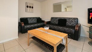 Un salon avec deux canapés à motifs, une table basse en bois, un meuble à bouteilles, un luminaire au plafond et un téléviseur posé sur un meuble.