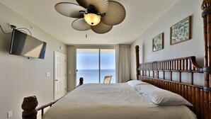 A bedroom with a bed, a ceiling fan, a TV mounted on the wall, and a view of the ocean through a door.