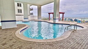 A swimming pool with a tiled deck, a covered area with chairs, and a view of the ocean.