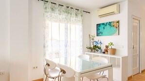 A dining area with a glass table and white chairs, a window with a bamboo curtain, a wall-mounted air conditioner, and a painting of a beach scene. - Marino house, your beach house (san francesc, formentera)