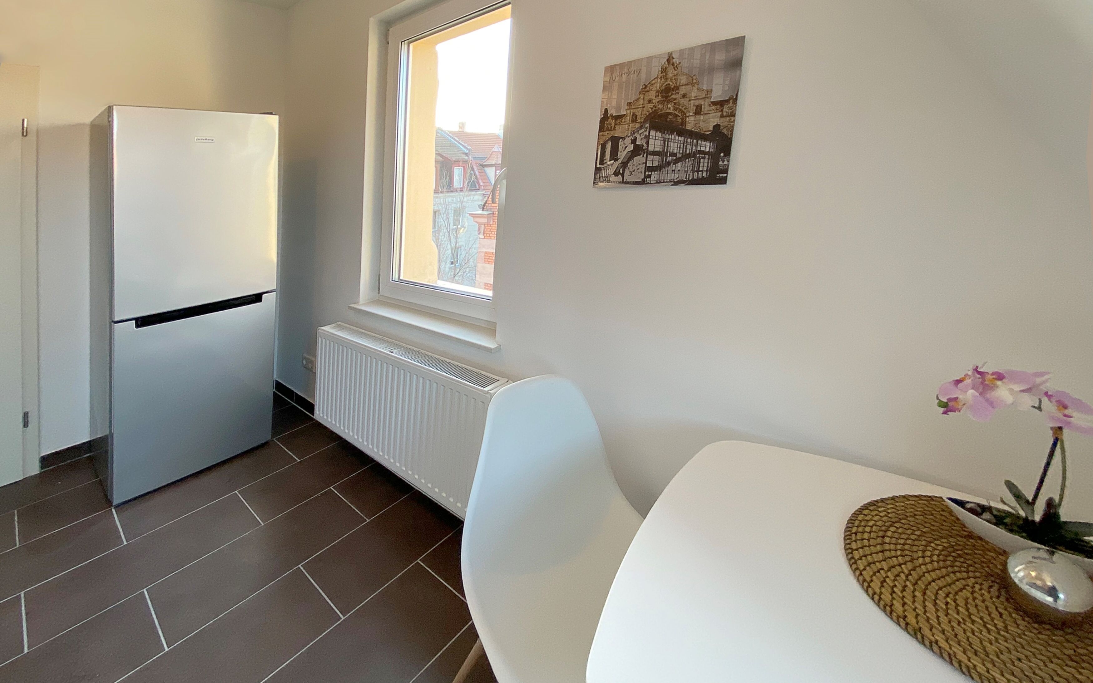 Une petite cuisine avec un réfrigérateur blanc, une fenêtre donnant sur des immeubles, un radiateur et une chaise blanche.