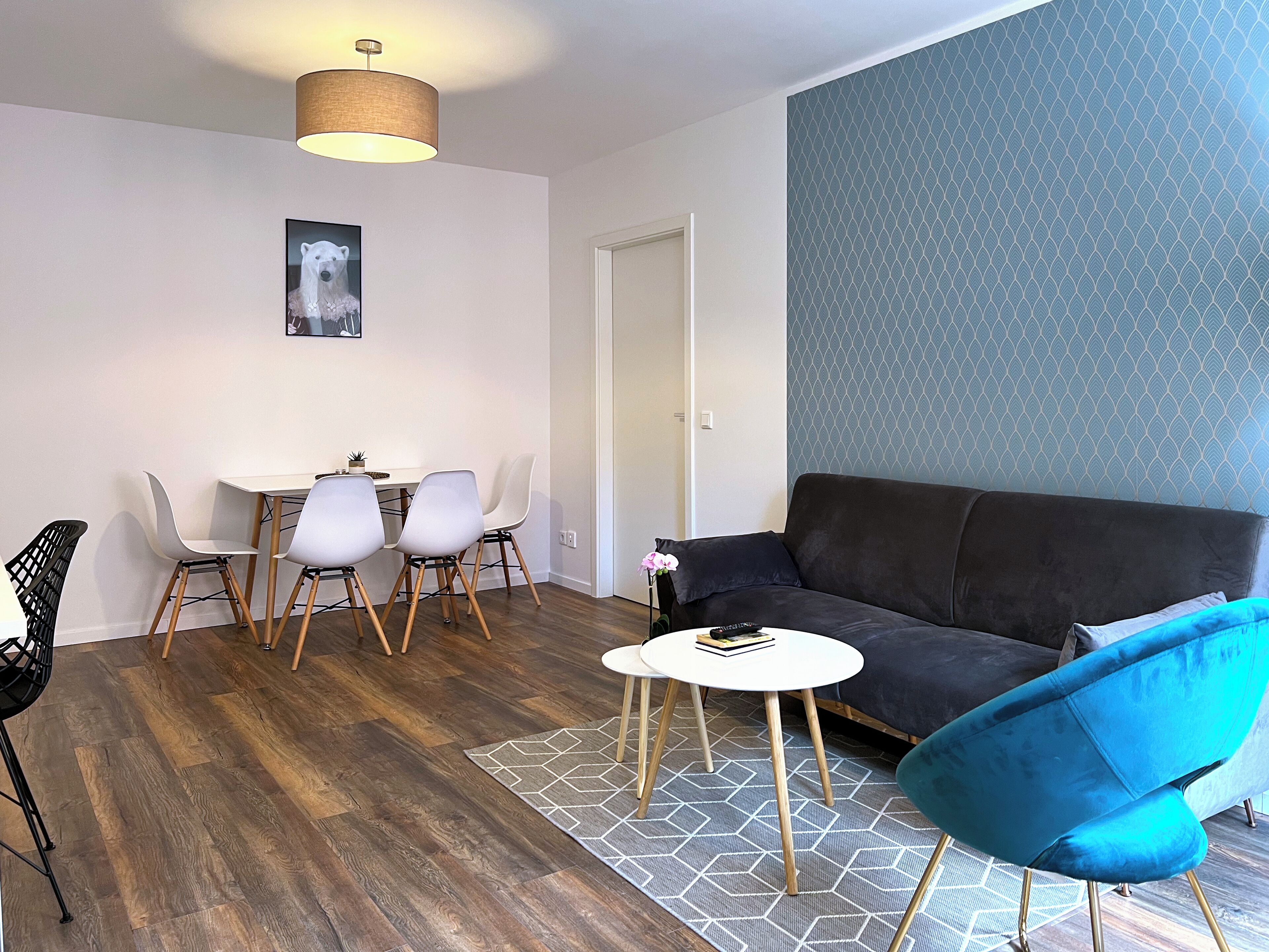 Una sala de estar moderna con un sofá marrón oscuro, un sillón azul, una mesa de centro blanca y un comedor con una mesa blanca y cuatro sillas.