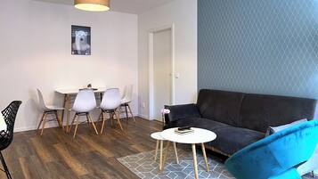 Una sala de estar moderna con un sofá marrón oscuro, un sillón azul, una mesa de centro blanca y un comedor con una mesa blanca y cuatro sillas.