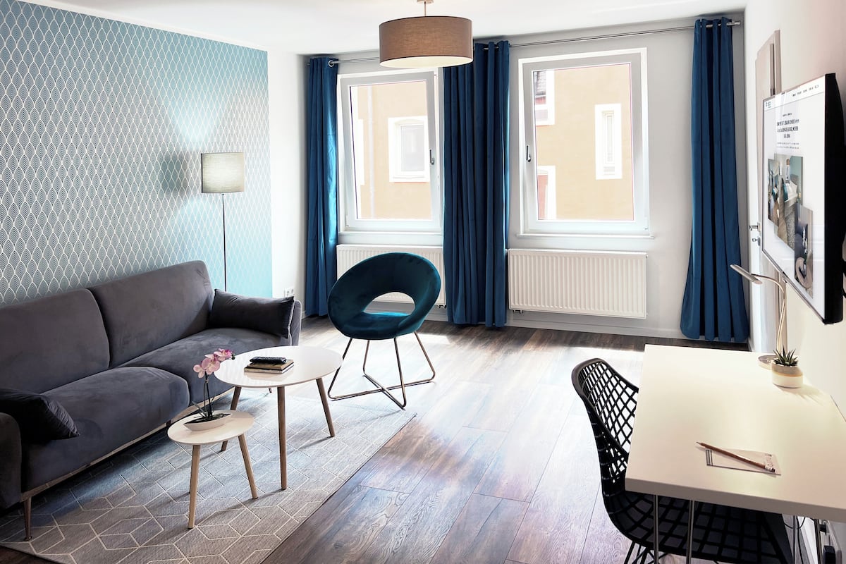 A modern living room with a sofa, a round table, and a chair. There are two windows with curtains, a radiator, and a lamp.