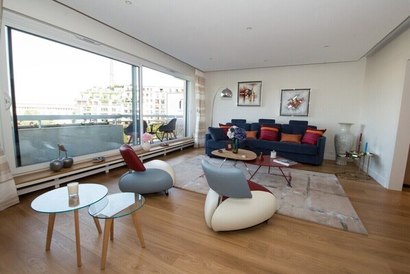 Ein modernes Wohnzimmer mit großem Fenster, einem blauen Sofa, einem Glastisch und einem roten Sessel.