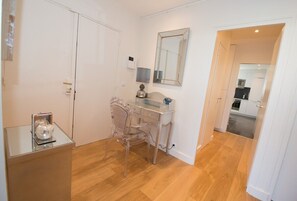 Ein Zimmer mit Schreibtisch, Stuhl und Spiegel an der Wand. Eine Tür führt in einen anderen Raum, ein kleiner Tisch mit einem dekorativen Gegenstand steht daneben.