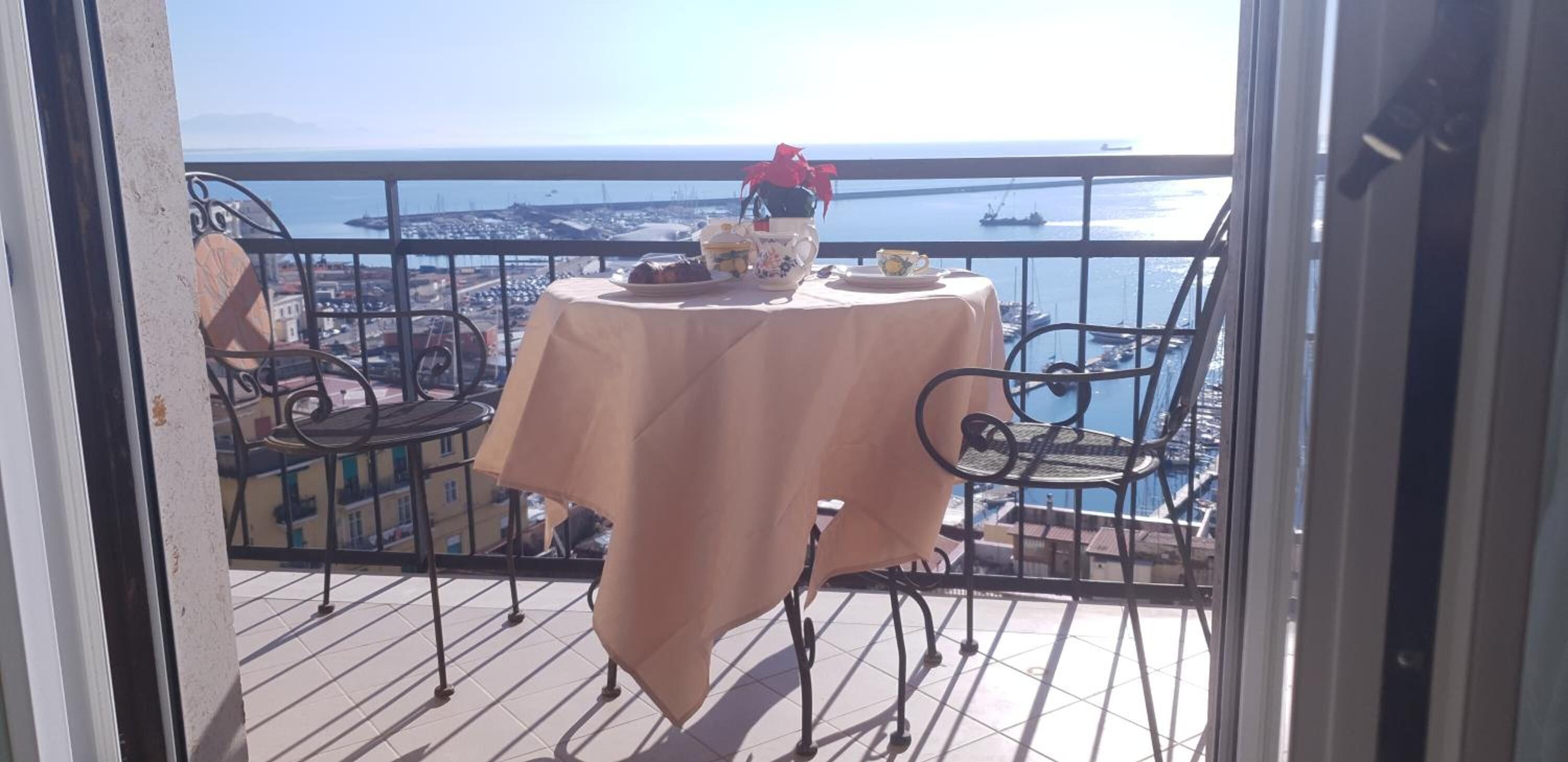 A balcony with a table set for two, overlooking a coastal cityscape with boats and a clear sky.