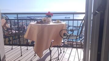 A balcony with a table set for two, overlooking a coastal cityscape with boats and a clear sky.