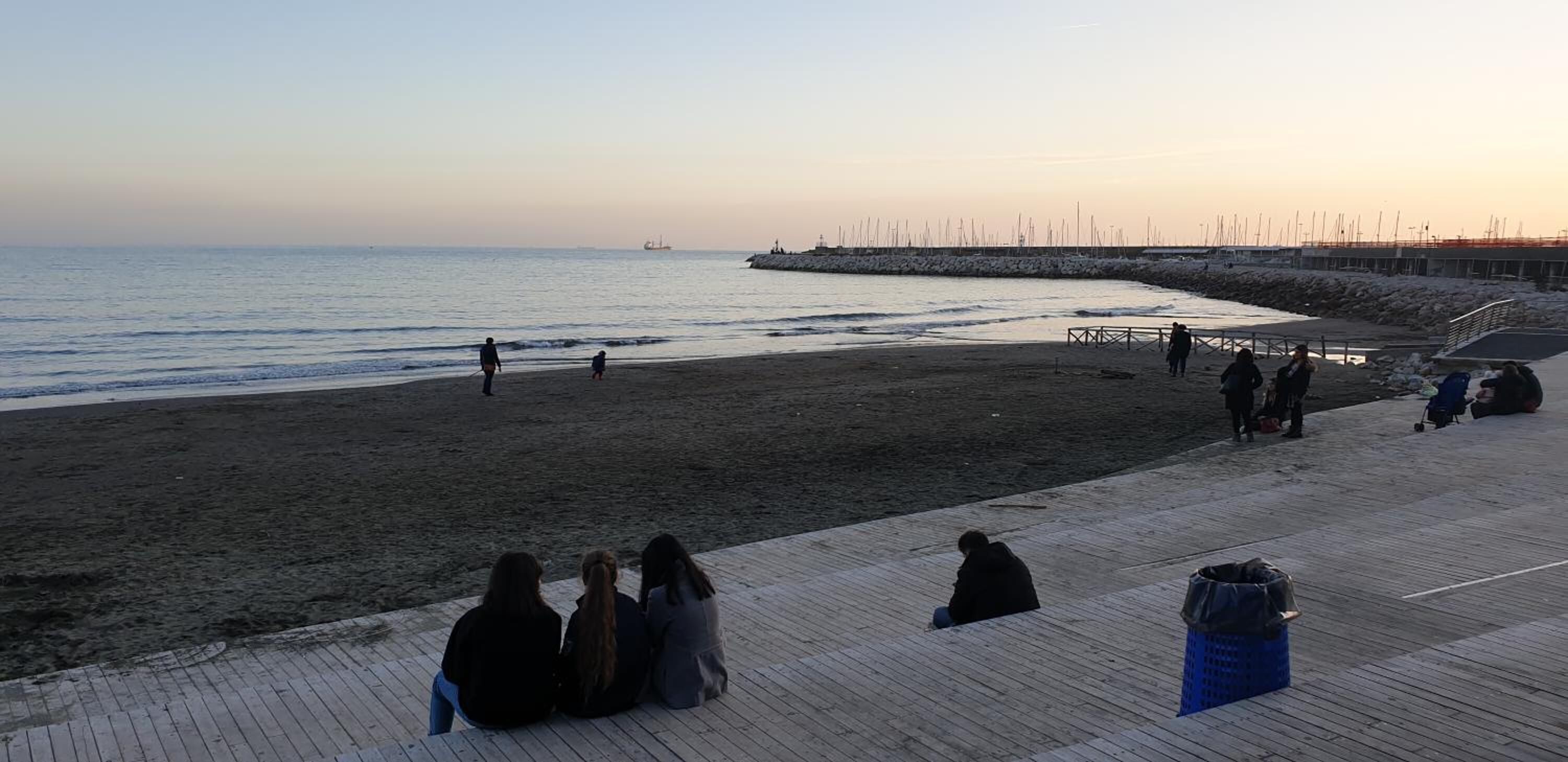 A beach with a wooden boardwalk, people walking and sitting, and a distant ship on the water.