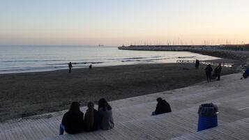 A beach with a wooden boardwalk, people walking and sitting, and a distant ship on the water.