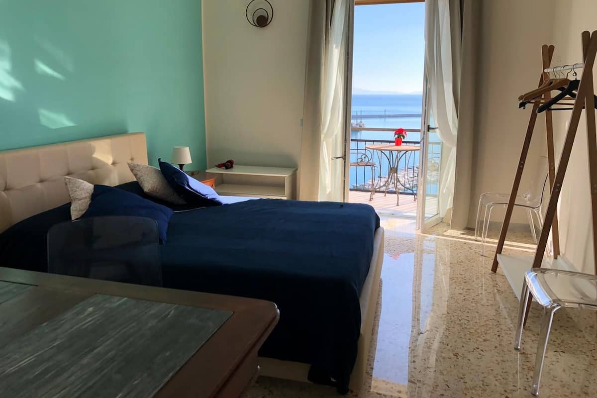 A hotel room with two beds, a small table, a chair, a balcony with a view of the sea, and a painting on the wall.