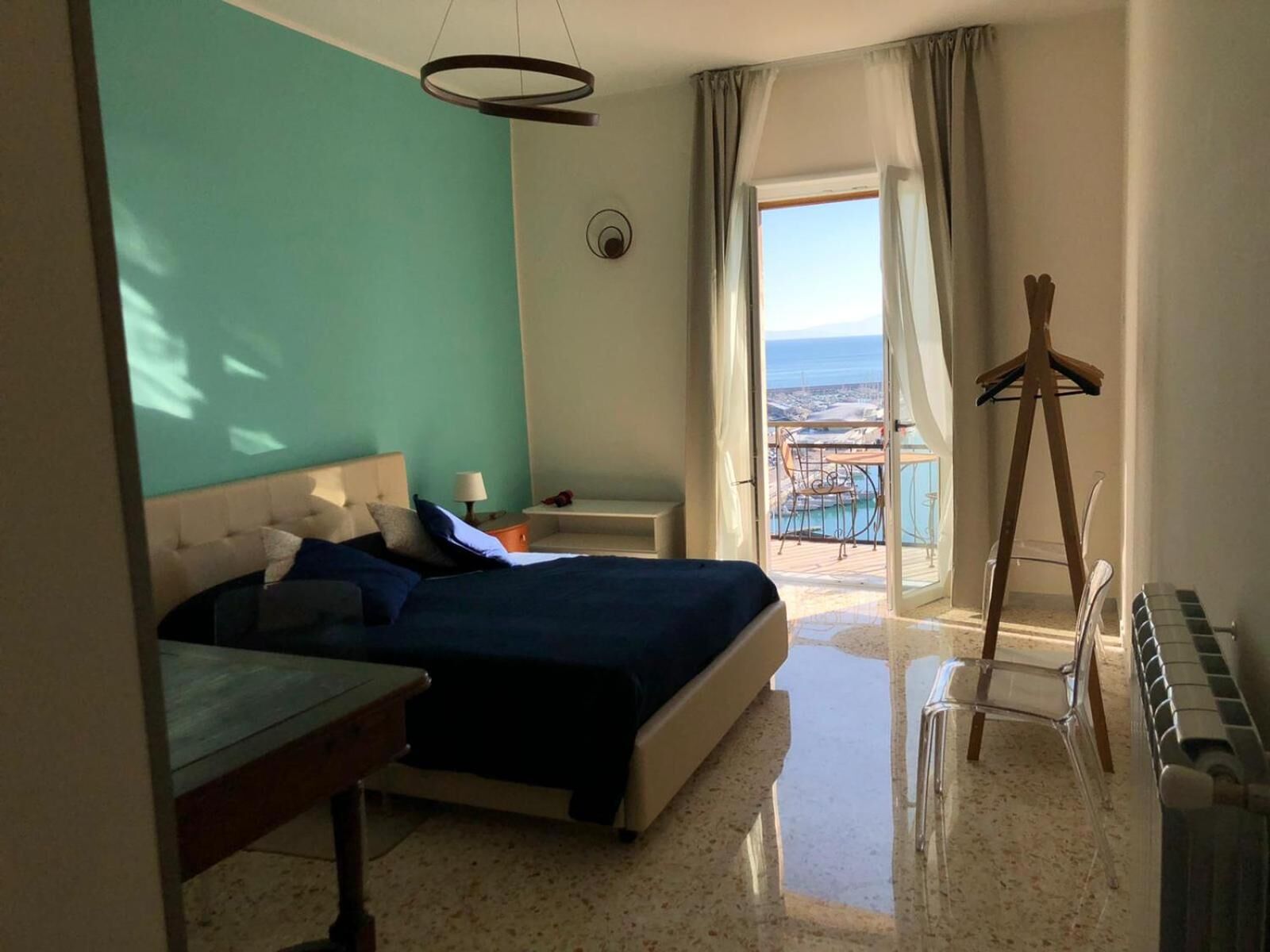 A hotel room with a bed, a desk, a chair, a radiator, and a view of the sea.