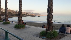 Eine Küstenpromenade mit Palmen, einer Holzpromenade und Blick auf das Meer sowie entfernte Gebäude.