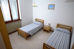 A small, clean room with two single beds, a bedside table with a lamp, a radiator, and a window with lace curtains. - Residence Fiore (Ledro)